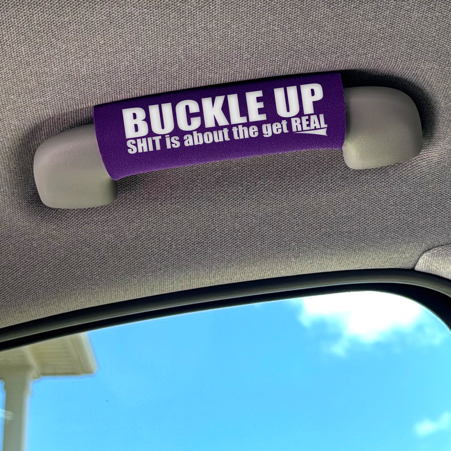 Car visor with a purple strap labeled 'BUCKLE UP SHIT is about the get REAL' against a blue sky background.