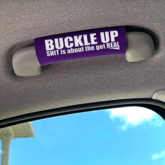 Car visor with a purple strap labeled 'BUCKLE UP SHIT is about the get REAL' against a blue sky background.